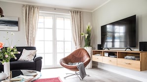 A modern living room with a leather chair, a flat-screen TV, a wooden TV stand, and a large window with curtains. - Ferienhaus Seeadler 7 - Der Natur Ganz nah (Fuhlendorf)
