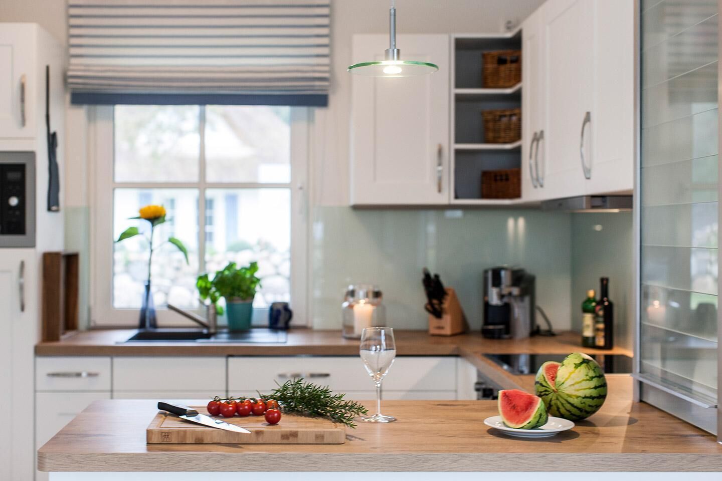 A modern kitchen with a wooden countertop, a cutting board with tomatoes, a glass of water, and a watermelon.
