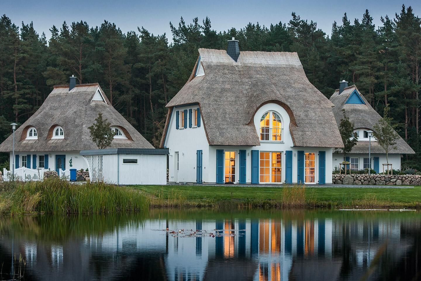 Eine Reihung aus reetgedeckten Häusern mit weißen Wänden und blauen Fensterläden, gelegen an einem stillen See mit Wald im Hintergrund.