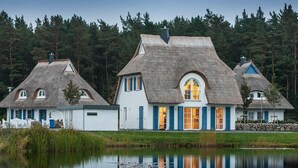 Una hilera de cabañas con techo de paja, paredes blancas y contraventanas azules, ubicadas junto a un lago tranquilo con un bosque al fondo.