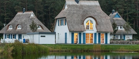 A row of thatched-roof cottages with white walls and blue shutters, situated by a calm lake with a forest backdrop.