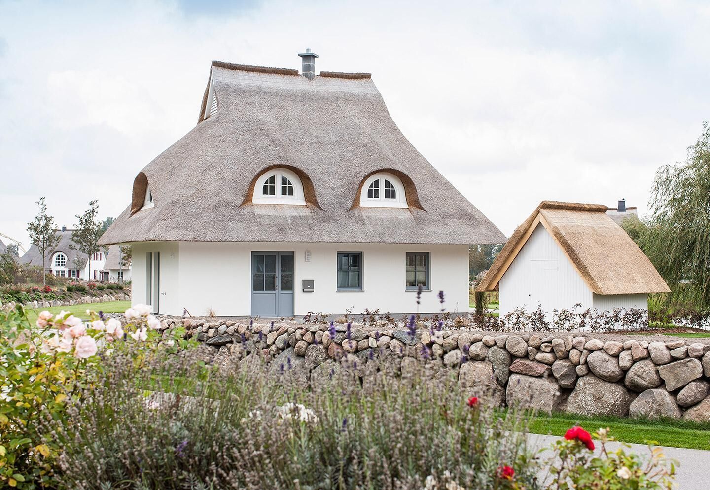Une maison traditionnelle à toit de chaume, dotée d’un mur en pierre à l’avant, et entourée d’un jardin où poussent diverses plantes et fleurs.