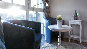Un moderno salón con un gran ventanal, una mesa redonda con una planta en maceta y una mesita auxiliar con una botella y vasos.