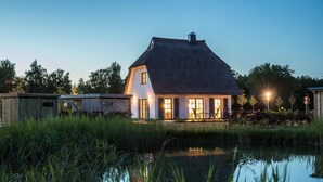 Una casa con tetto di paglia e pareti bianche, immersa in un'acqua calma con erba alta in primo piano.
