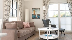 Un soggiorno con un divano beige, un tavolino bianco rotondo e una sedia. Appeso al muro, un quadro raffigura un faro.