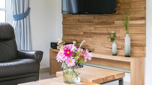 A modern living room with a wooden wall, a flat-screen TV, a glass coffee table, a leather sofa, and a vase with flowers. - Ferienhaus Haubenlerche 16 - Der Natur Ganz nah (Fuhlendorf)