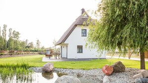 A white house with a thatched roof, a pond, and a garden with rocks and trees. - Ferienhaus Seeschwalbe 37 - Der Natur Ganz nah (Fuhlendorf)