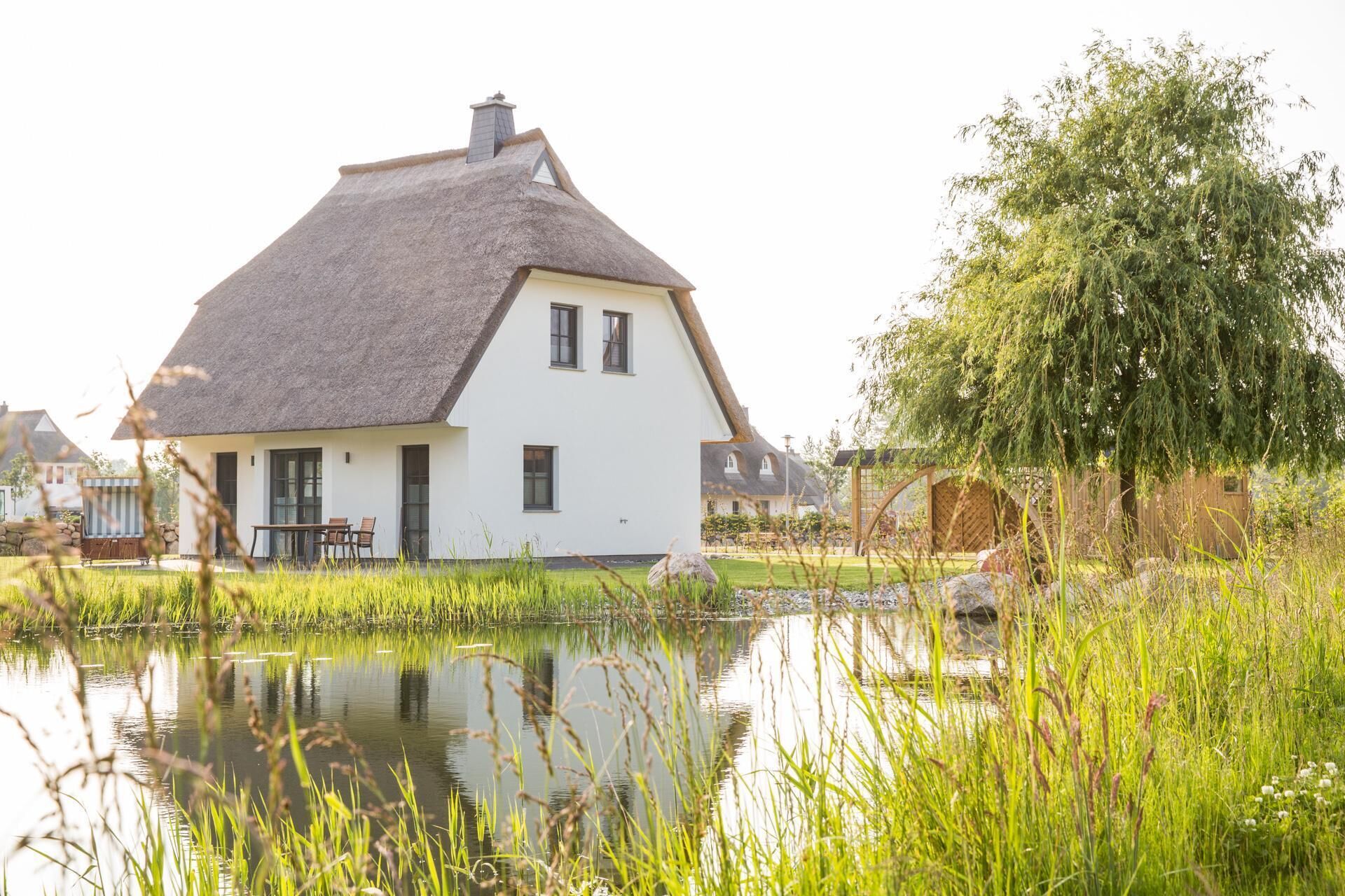 A white house with a thatched roof, a pond in the foreground, and a tree to the right.