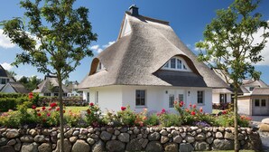 Ein Haus mit Strohdach und einer Steinsäule, umgeben von einem Garten mit verschiedenen Blumen.
