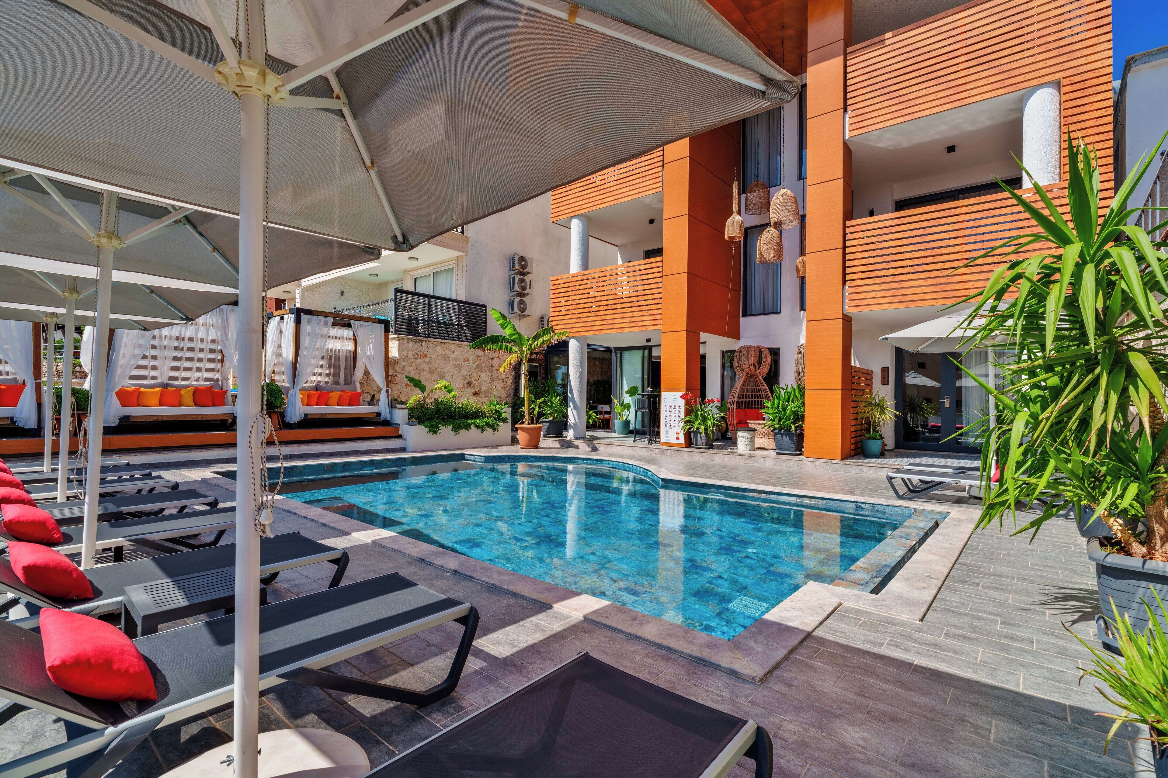 Un área de piscina con sillones de descanso y sombrillas, rodeada por un edificio moderno con pared de piedra y detalles de madera.