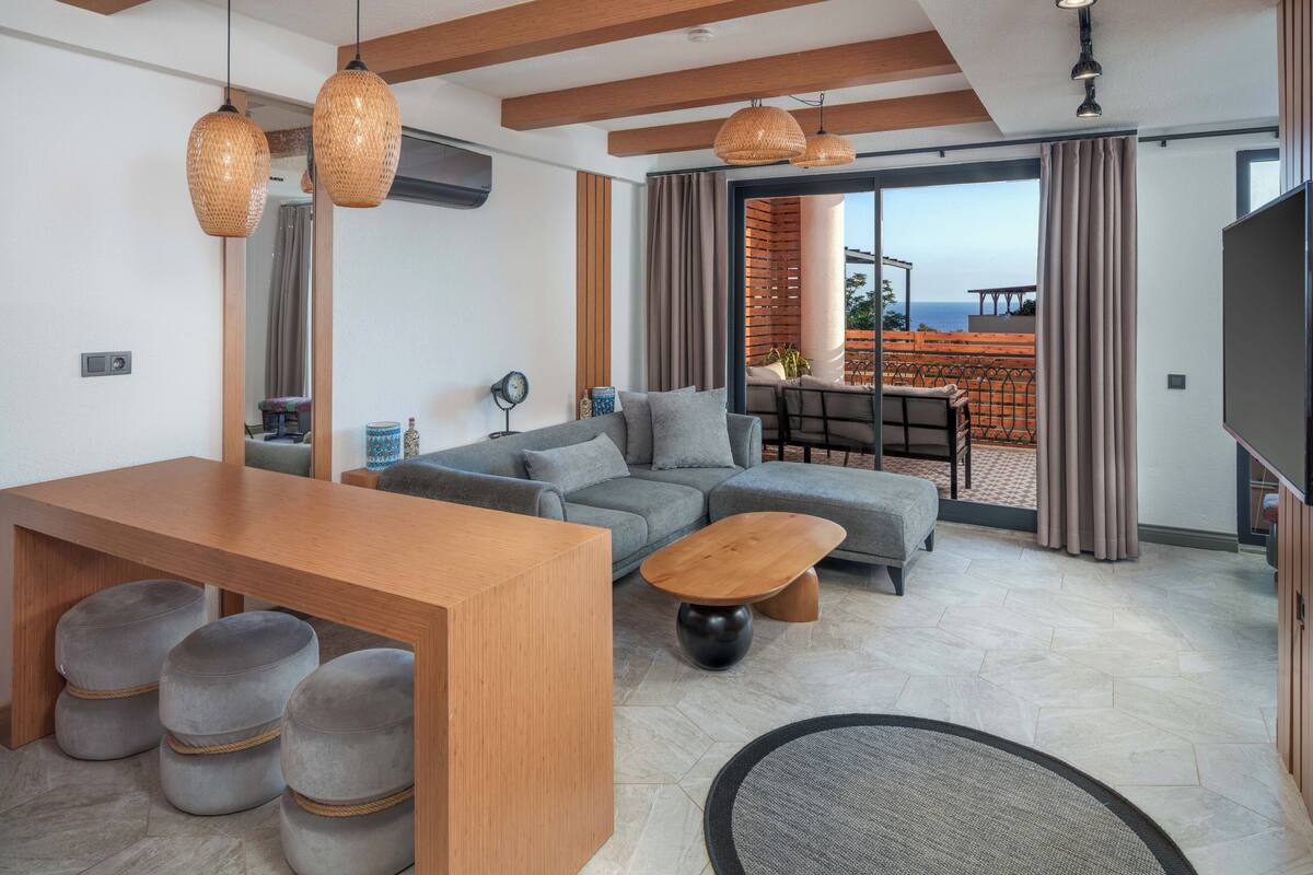 A modern living room with a wooden dining table, grey sofas, and a large window with a view of the sea.