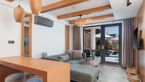 Una sala de estar moderna con sofá, reposapiés y una mesa de comedor de madera. Hay lámparas de mimbre, un televisor de pantalla plana y un ventanal corredero que da a un área exterior con piscina.