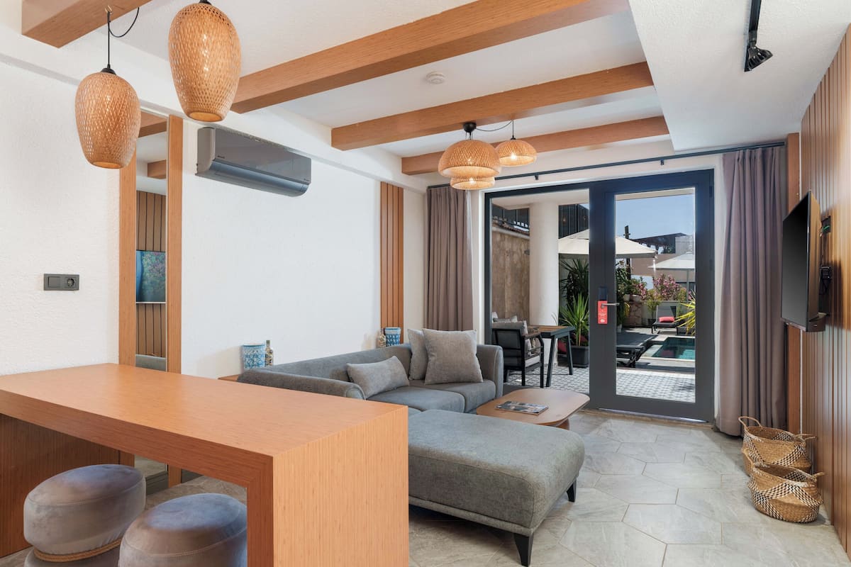 A modern living room with a sofa, ottoman, and a wooden dining table. There are wicker lamps, a flat-screen TV, and a sliding glass door leading to an outdoor area with a pool.