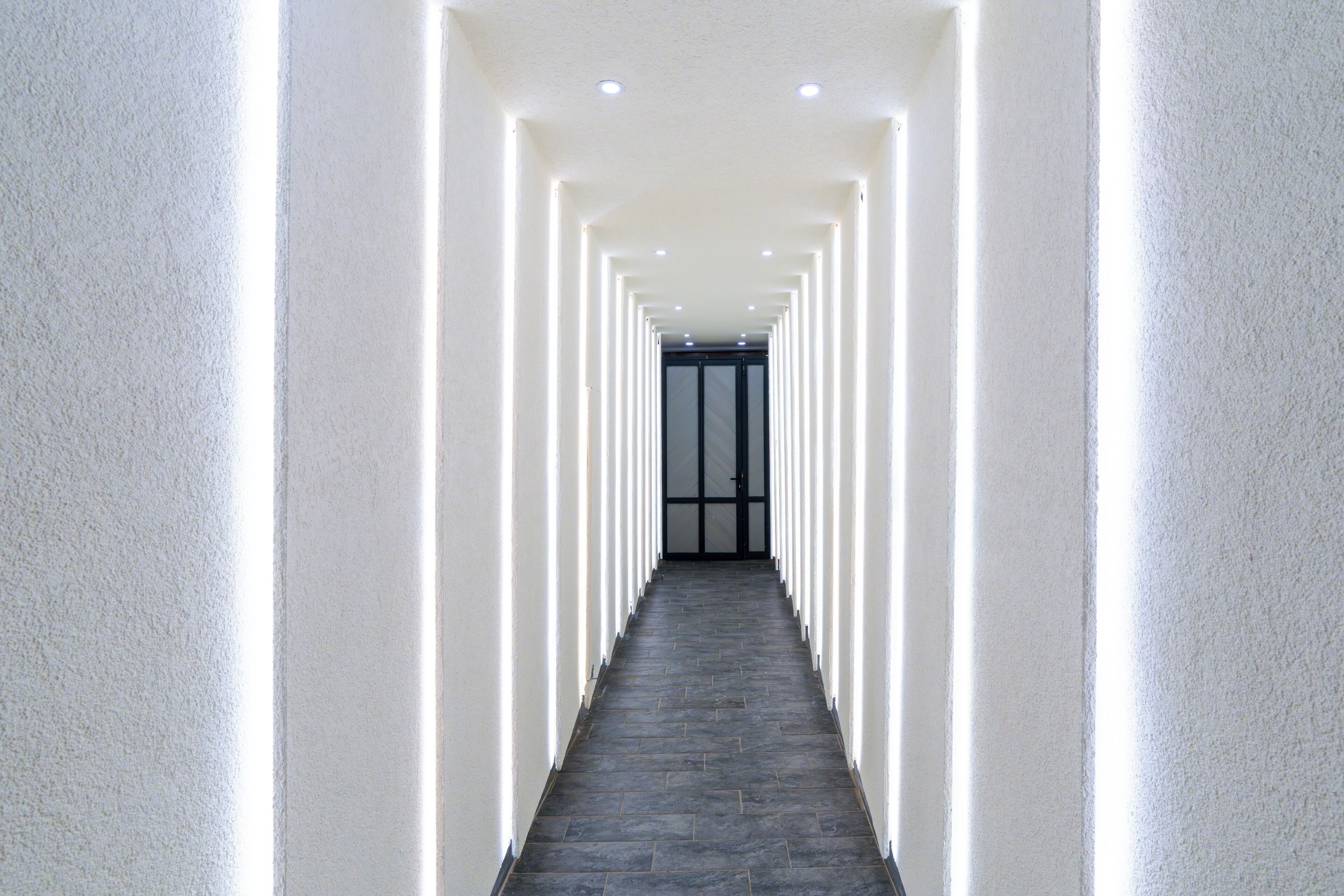 Un long couloir étroit aux murs blancs et au sol sombre, qui mène à une porte au fond.