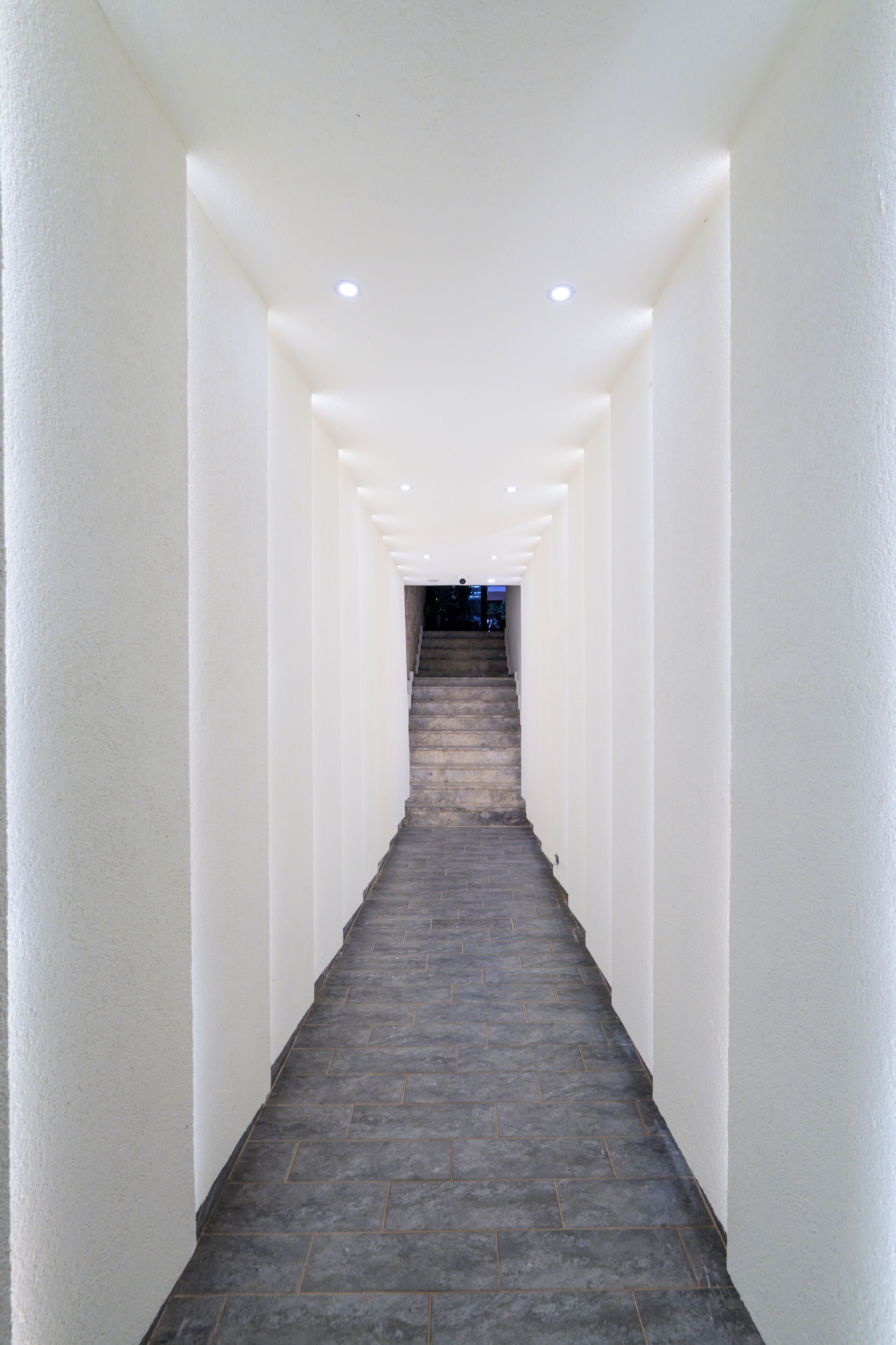 Un pasillo estrecho y bien iluminado, con paredes blancas y un suelo de baldosas oscuras que conduce a una escalera.