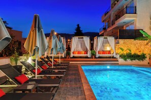 Zona de piscina con tumbonas y sombrillas, una piscina cristalina y un edificio con balcones.
