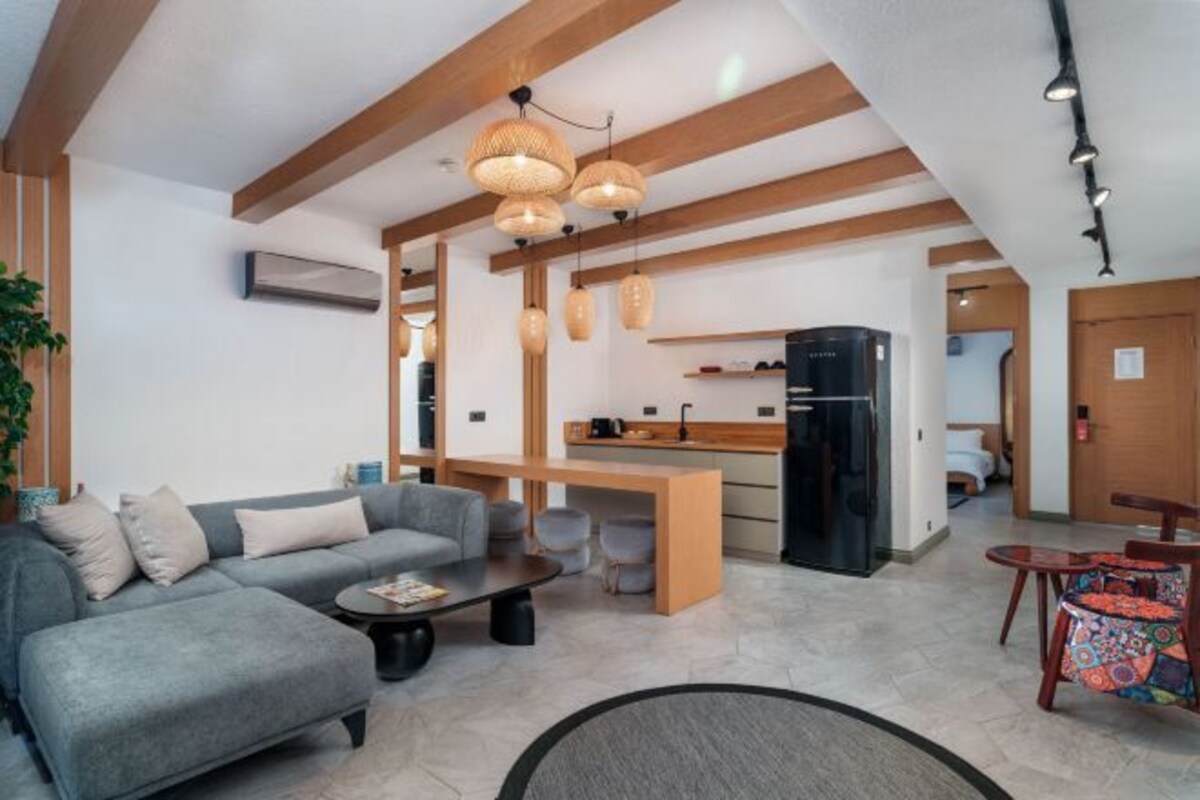 Una sala de estar moderna con un sofá gris, una mesa de centro y una zona de comedor con mantel colorido. Hay una cocina con un refrigerador negro y vigas de madera en el techo.