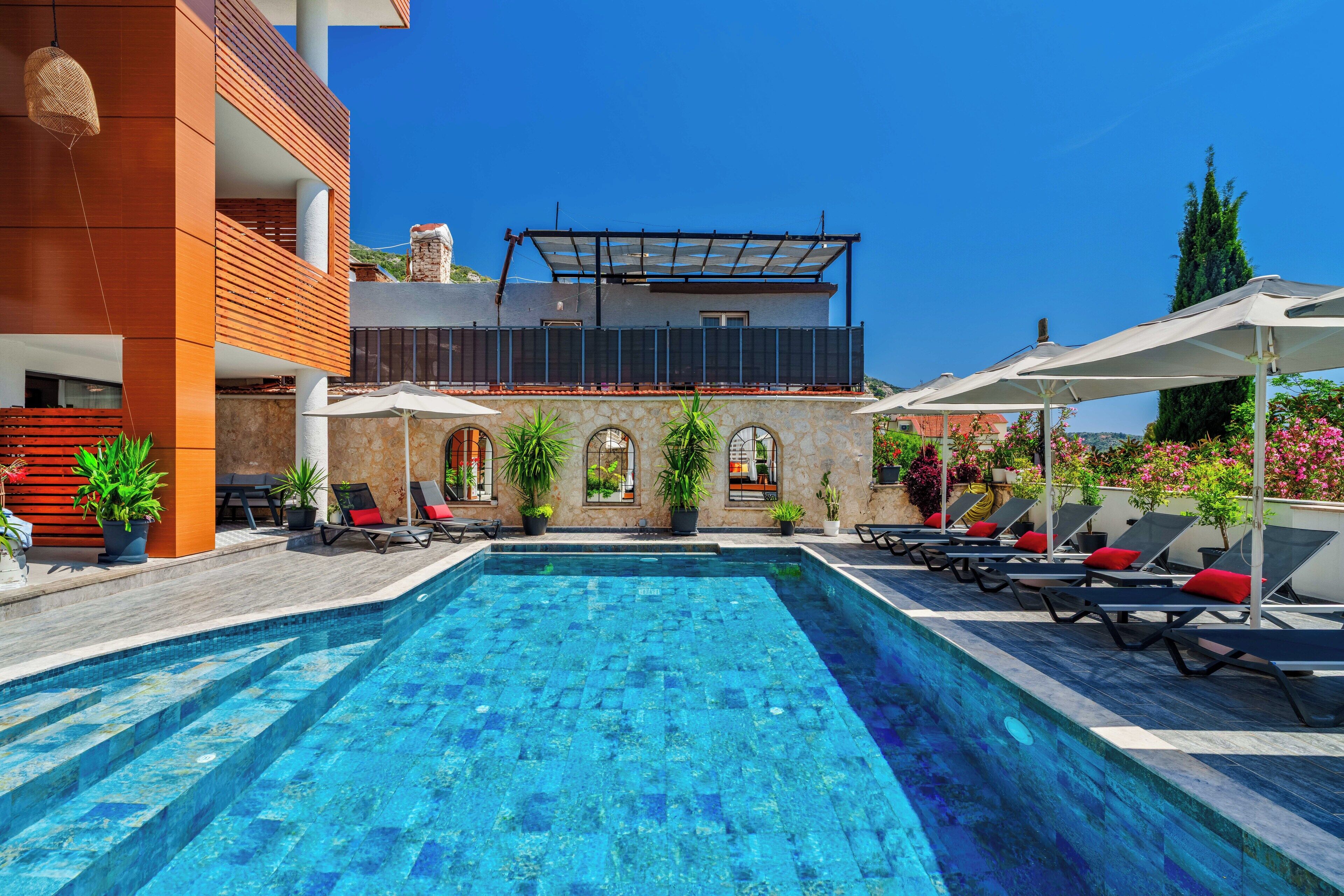 A swimming pool with sun loungers and umbrellas, surrounded by potted plants and a modern building with a balcony.