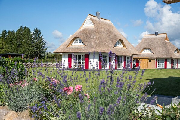 A thatched-roof house with red shutters, surrounded by a well-maintained garden with purple and pink flowers.