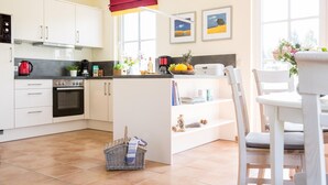A modern kitchen with white cabinets, a built-in oven, and a dining area with a white table and chairs. - Ferienhaus Schilfrohrsänger 48 - Der Natur Ganz nah (Fuhlendorf)