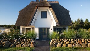 Et traditionelt bindingsværkshus med stråtækt tag, en midterdør og vinduer, omgivet af en stenvæg og grøn vegetation.