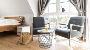 Een moderne hotelkamer met een houten bed, twee grijze fauteuils, een klein rond tafeltje met een drankje en een groot raam met gordijnen.