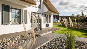 Een modern huis met rieten dak, grote ramen met luiken, en een goed onderhouden tuin met stenen en planten.