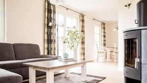 A modern living room with a sofa, a coffee table, a fireplace, and a dining area in the background. - Ferienhaus Schilfrohrsänger 30 - Der Natur Ganz nah (Fuhlendorf)