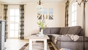 A living room with a grey sofa, a coffee table, a bookshelf, and a large window with curtains. - Ferienhaus Schilfrohrsänger 30 - Der Natur Ganz nah (Fuhlendorf)