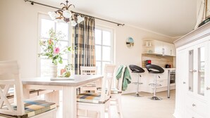 A bright kitchen with a dining table, chairs, and two bar stools. There is a window with curtains, a chandelier, and a cabinet with a stove. - Ferienhaus Schilfrohrsänger 30 - Der Natur Ganz nah (Fuhlendorf)