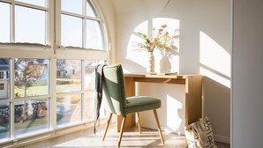 A room with a large window, a desk with a vase of flowers, a chair, and a bag on the floor. - Ferienhaus Schilfrohrsänger 36 - Der Natur Ganz nah (Fuhlendorf)