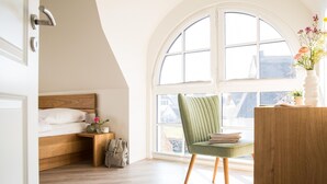 A bedroom with a wooden bed, a green chair, a desk with books, and a large arched window. - Ferienhaus Schilfrohrsänger 36 - Der Natur Ganz nah (Fuhlendorf)