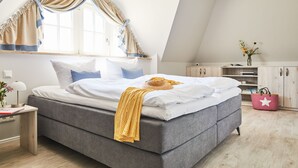 Ein modernes Schlafzimmer mit einem grauen Bett, weißer Bettwäsche und einer gelben Decke. Es gibt ein Fenster mit Vorhängen, einen Nachttisch mit einer Lampe und ein kleines Regal mit einer pinkfarbenen Tasche.