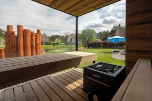 A wooden deck with a bench, a Harvia fire pit, and a view of a garden and houses.