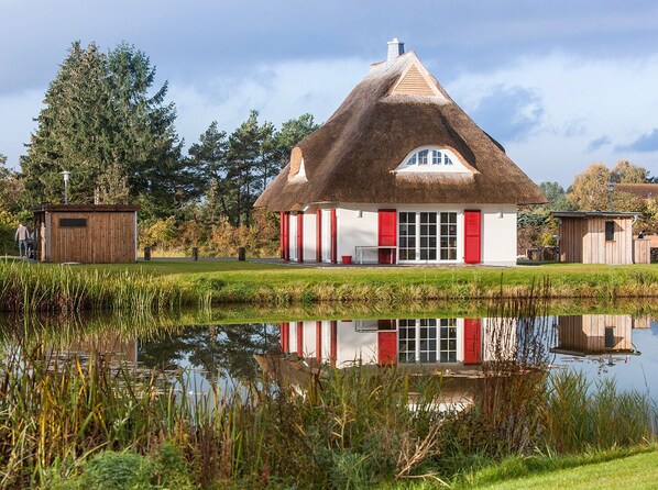 A thatched-roof house with a white facade and red shutters, situated beside a calm pond with a clear reflection.