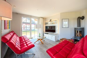 Ein modernes Wohnzimmer mit roten Ledersofas, einem Glastisch, einem Flachbildfernseher, einem Kaminofen und einem großen Fenster mit Blick auf ein Gebäude.