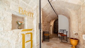 A stone-walled corridor with a large golden 'B' sign, a vase with flowers, a metal chair, and a golden urn. - Bayit Charming Place (Polignano a Mare)