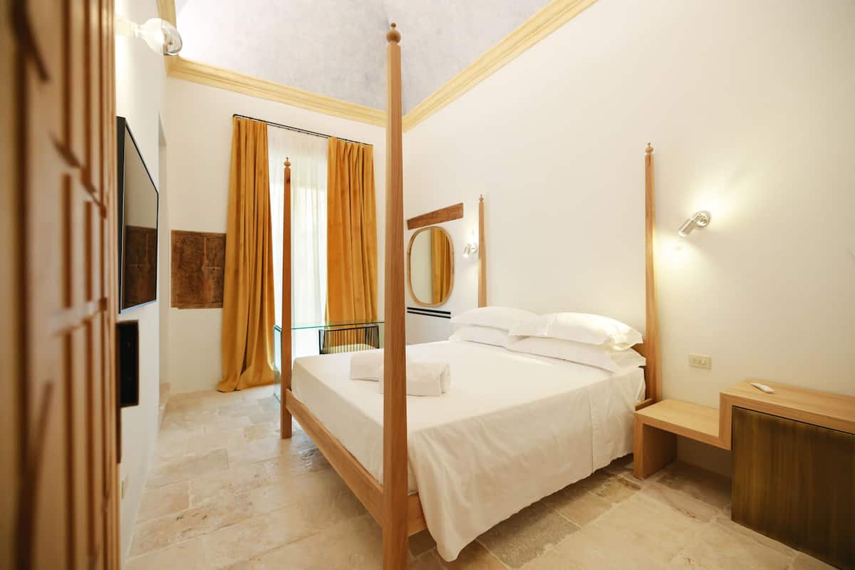 A four-poster bed with white linens, a bedside table, a mirror, and a wall-mounted light fixture.