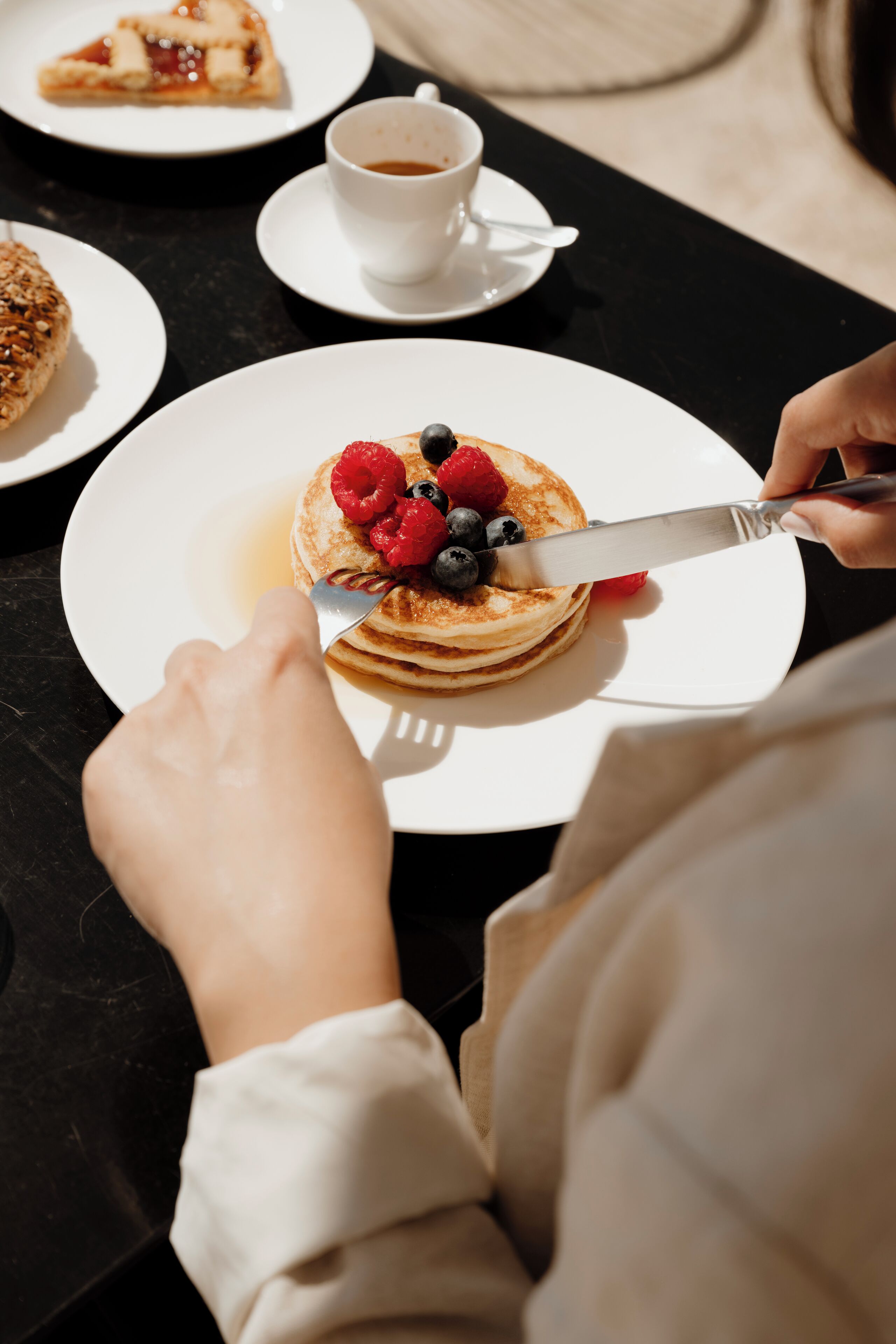 Een ontbijtsetting met pannenkoeken belegd met bessen en siroop, een kop koffie en een plakje taart op een tafel.