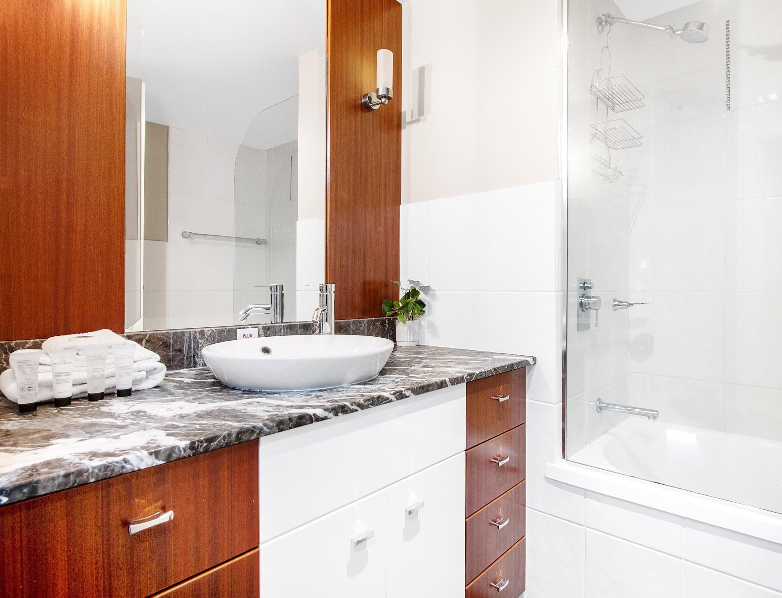 Un baño moderno con un lavabo blanco, encimera de mármol, armarios de madera y cabina de ducha de vidrio.
