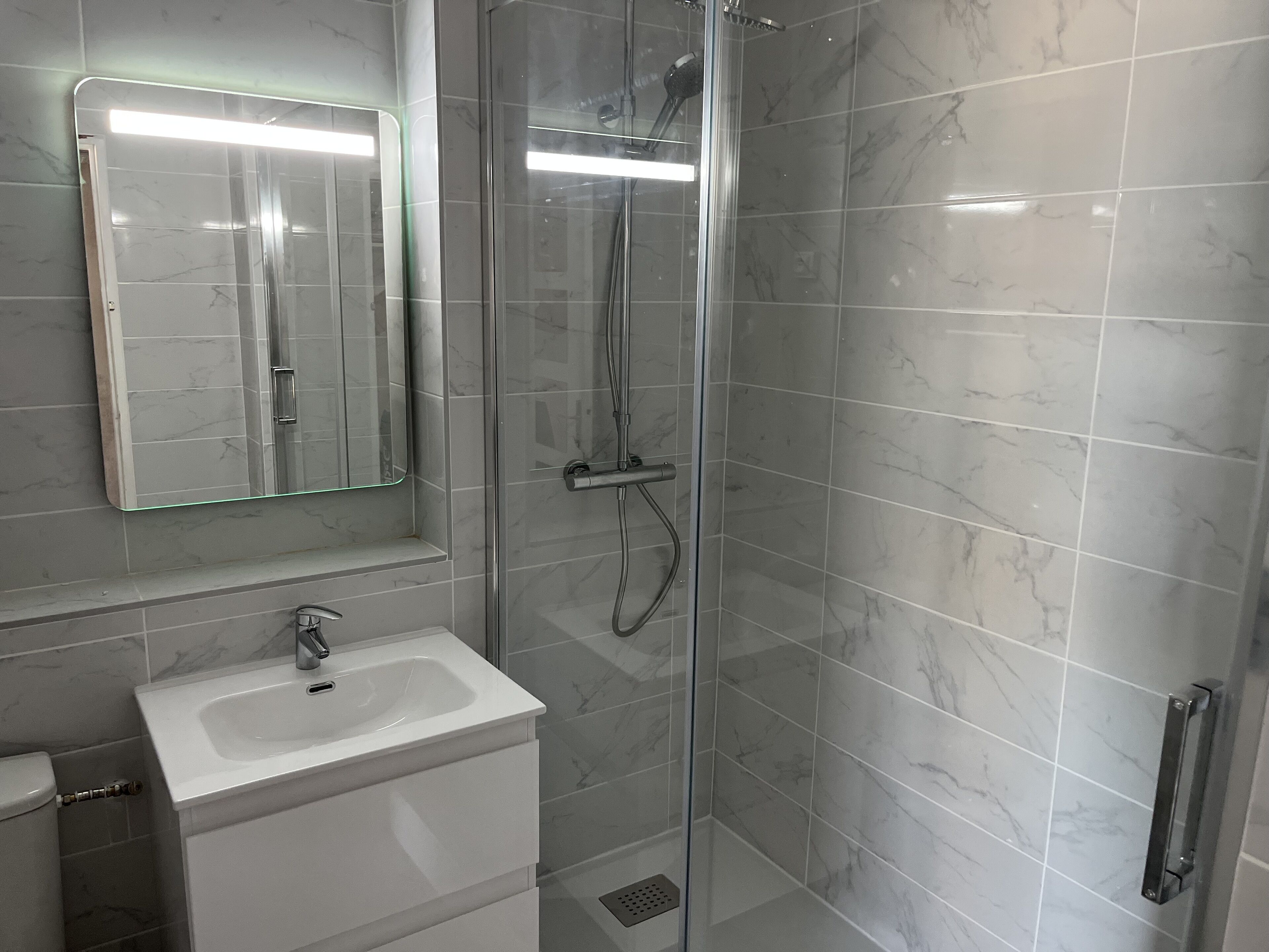 Une salle de bain avec une douche à parois de verre, un lavabo blanc et un miroir avec un éclairage au-dessus.