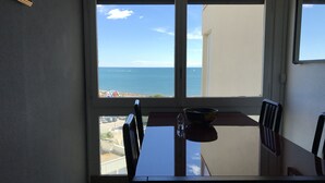 A dining table with chairs, a bowl, and a bottle of water in front of a window with a view of the sea and a beach. - VIEW OF THE MEDITERRANEAN SEA (La Grande-Motte)