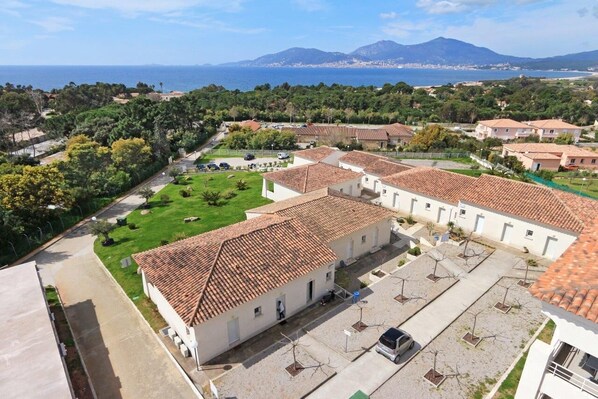 Un quartier résidentiel avec des bâtiments aux toits de tuiles en terre cuite, une pelouse verdoyante et une vue sur la mer et les montagnes en arrière-plan.