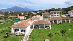A residential area with multiple two-story houses with tiled roofs, surrounded by greenery and a mountain in the background.