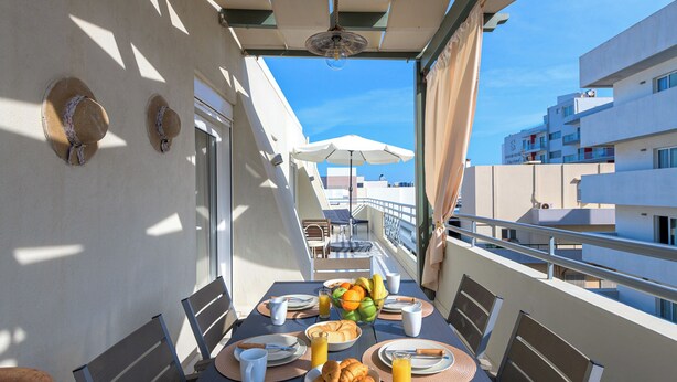 Un balcón con una mesa dispuesta para el desayuno, que incluye croissants, fruta y jugo, con vistas a un paisaje urbano.