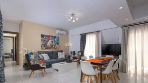 Una sala de estar moderna con sofá, mesa de centro, mesa de comedor, sillas, televisor y un cuadro de colores en la pared.