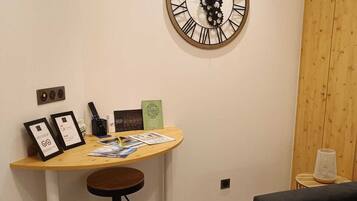 A modern living room with a round wall clock, a small wooden table with a stool, a black sofa, and a small side table with a lamp.