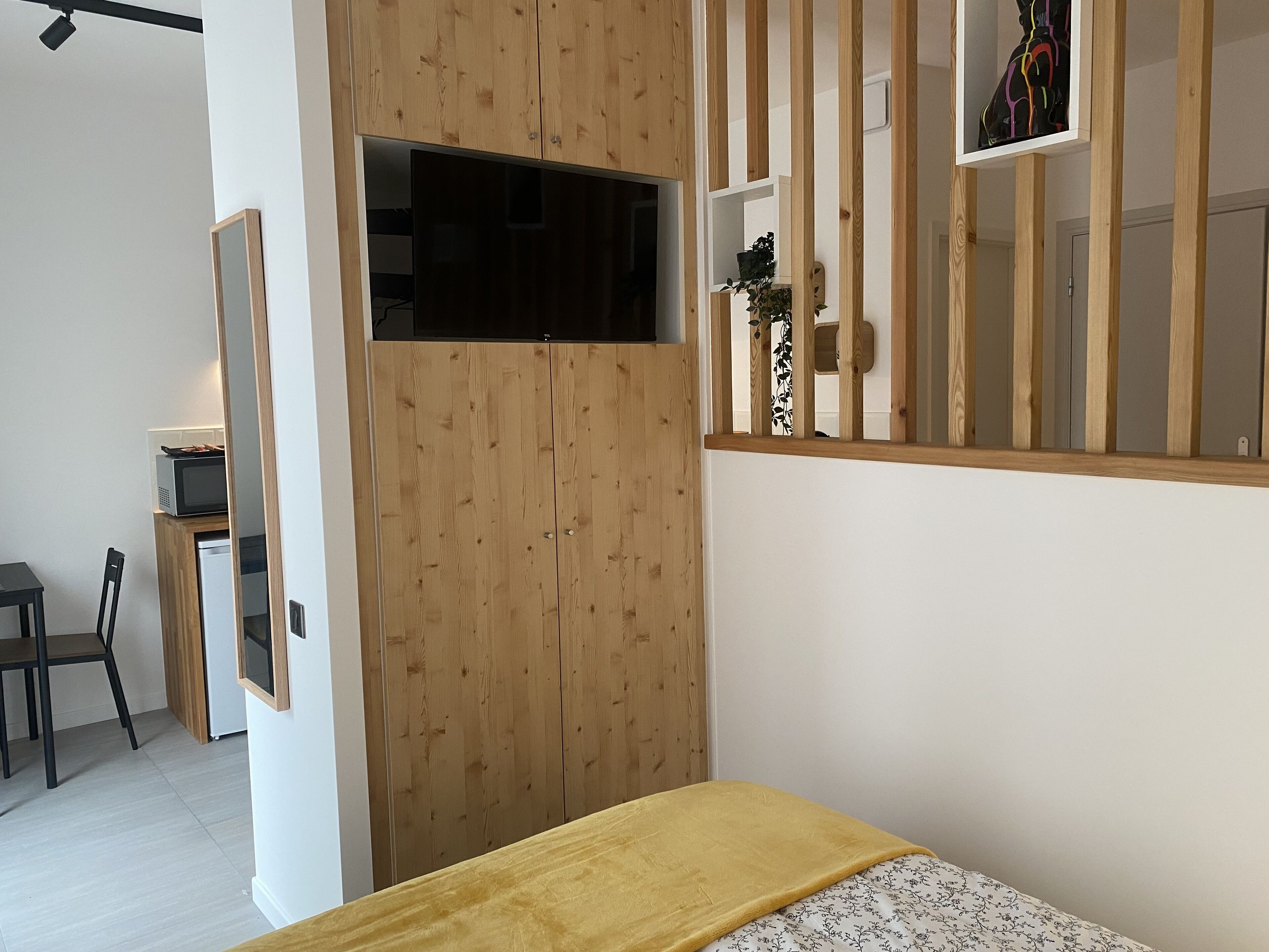 Een moderne slaapkamer met een houten garderobe, een bed met een gele deken, een klein bureau met een computer en een raam met een plant.