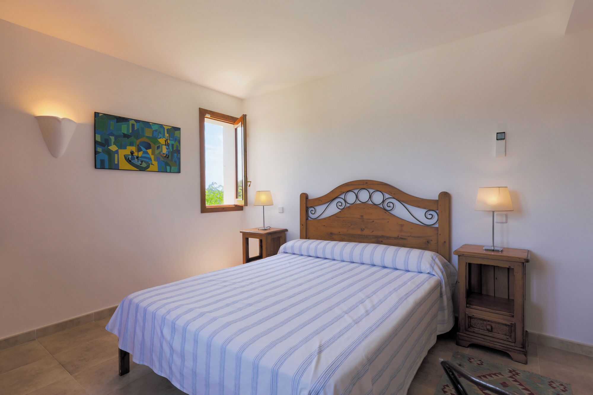 Une chambre à coucher comprenant un lit, deux tables de chevet, une commode, une lampe fixée au mur, un tableau et une fenêtre.
