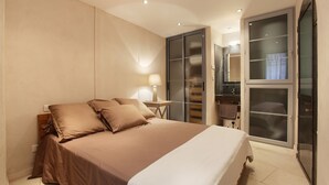 Un dormitorio con una cama grande, una mesita de noche con lámpara, un armario con puerta corrediza de vidrio y un baño con espejo y perchero.